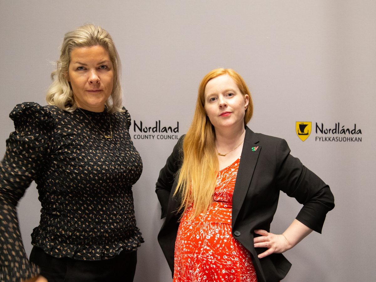 Aase Refsnes og Åshild Pettersen står foran en hvit bakgrunn som har logoen til Nordland fylkeskommune. Bilde