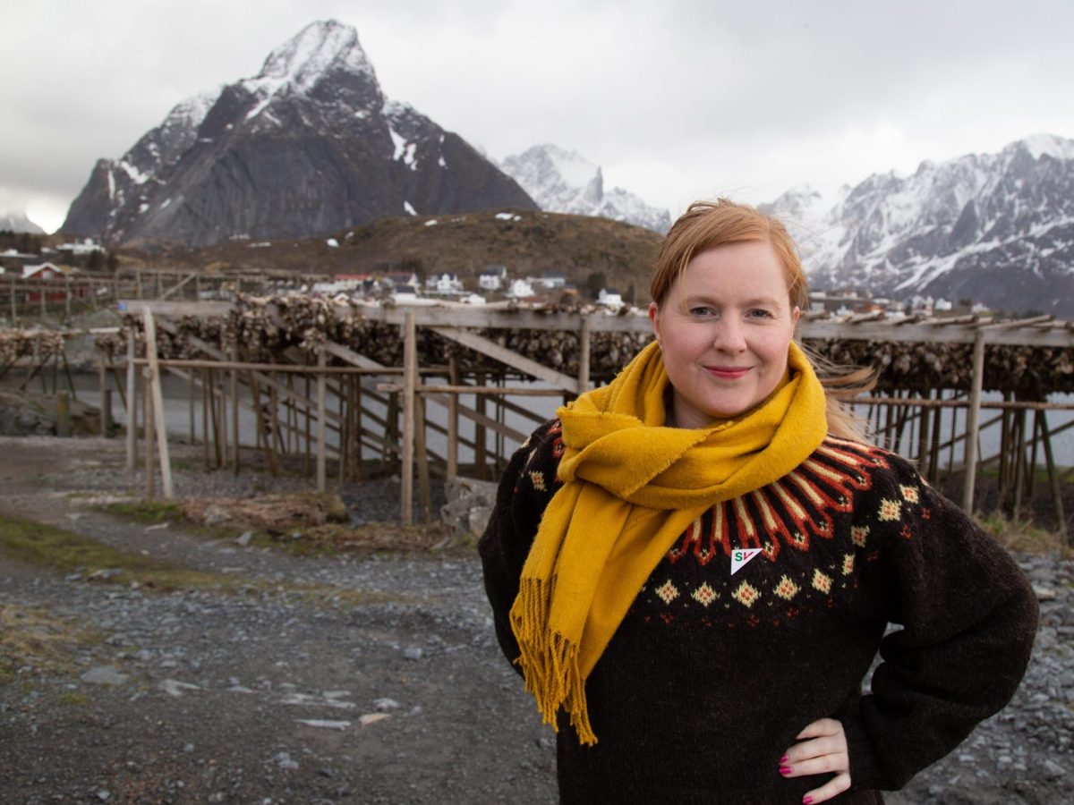 ÅShild Pettersen står foran en fiskehjell på Reine. Hun har henden i siden. I bakgunnen er det fjell. Bilde.