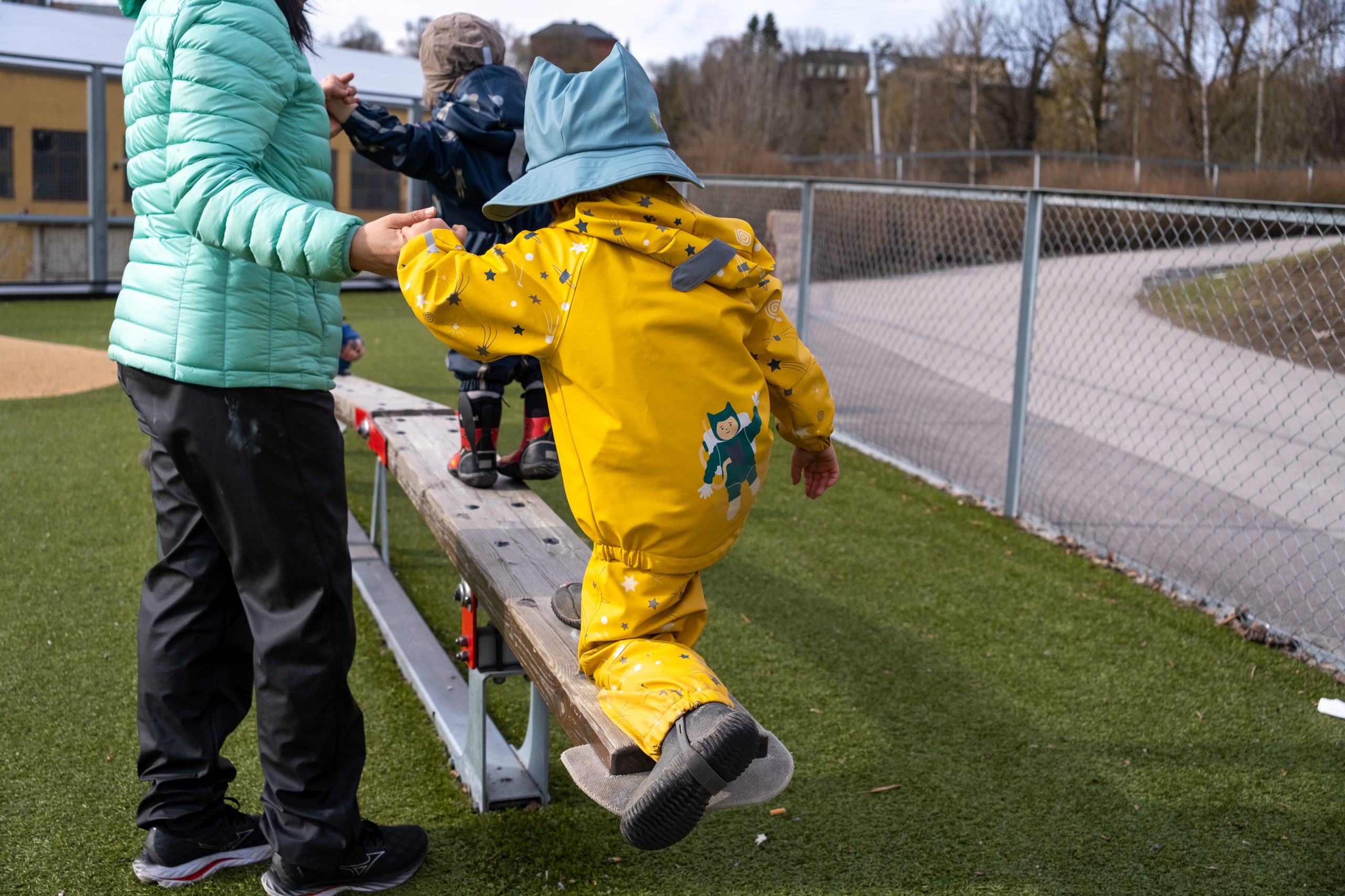 En kvinne holder to barn i hånda mens de balanserer på et lekeapparat, en slags opphøyd planke. Man ser menneskene kun bakfra. Bilde.