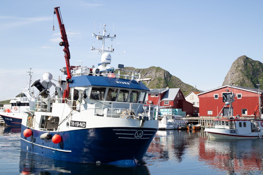 Bilde av en fiskebåt å vei til kai i Værøy. Fjell og røde hus i bakgrunnen.