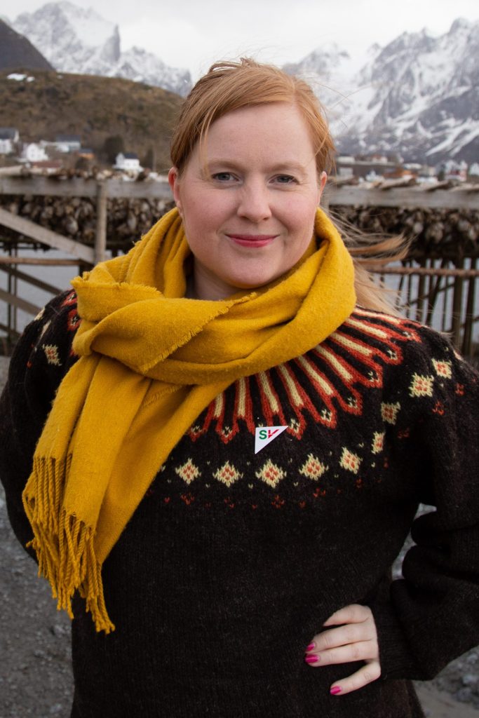 Åshild Pettersen står foran en fiskehjell på Reine. Hun har henden i siden. Hun har på seg ullgenser og et ullskjerf.I bakgunnen er det fjell. Bilde.