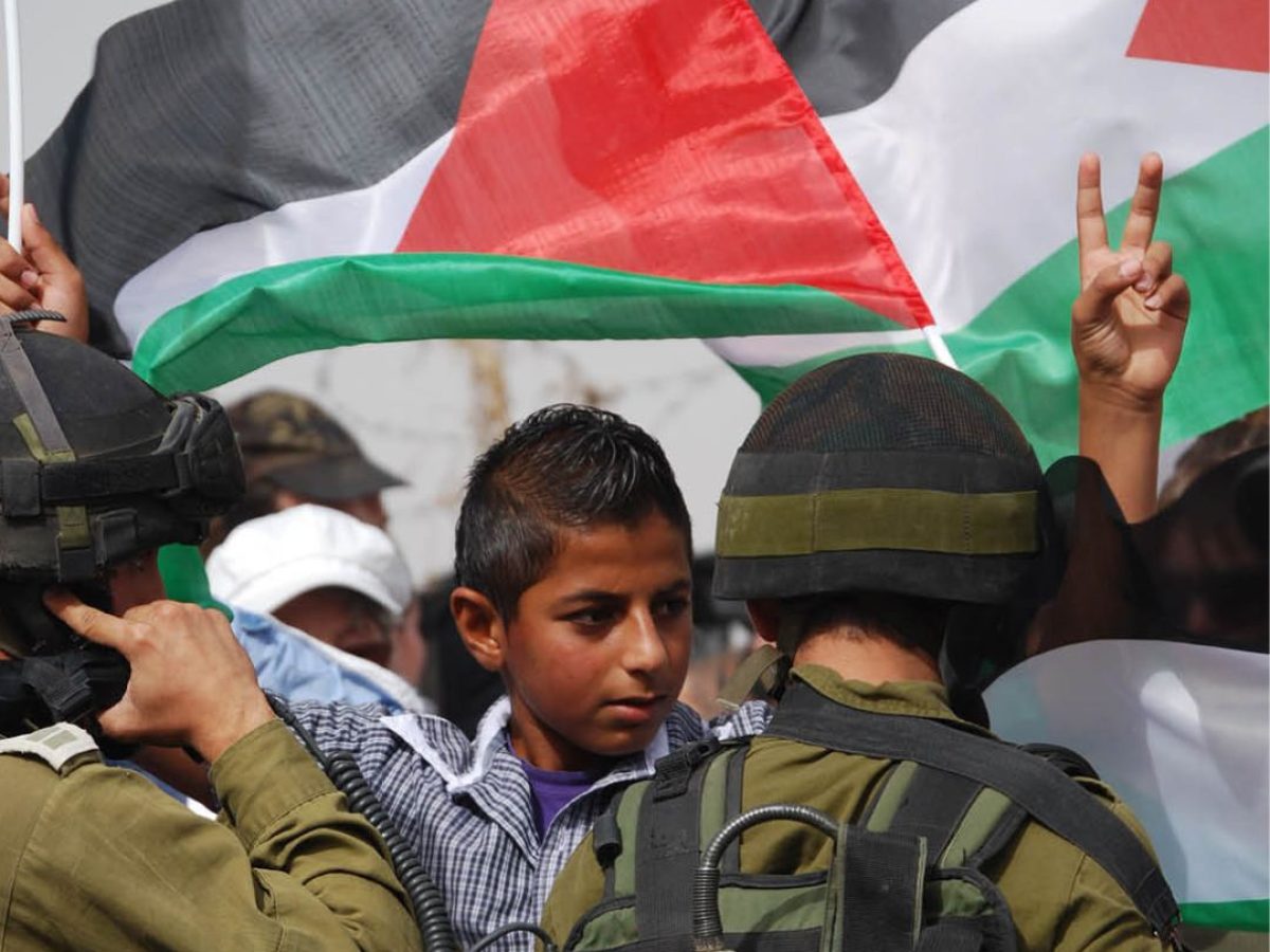 Palestinsk gutt foran palestinsk flagg gjør fredstegn til Israelske soldater