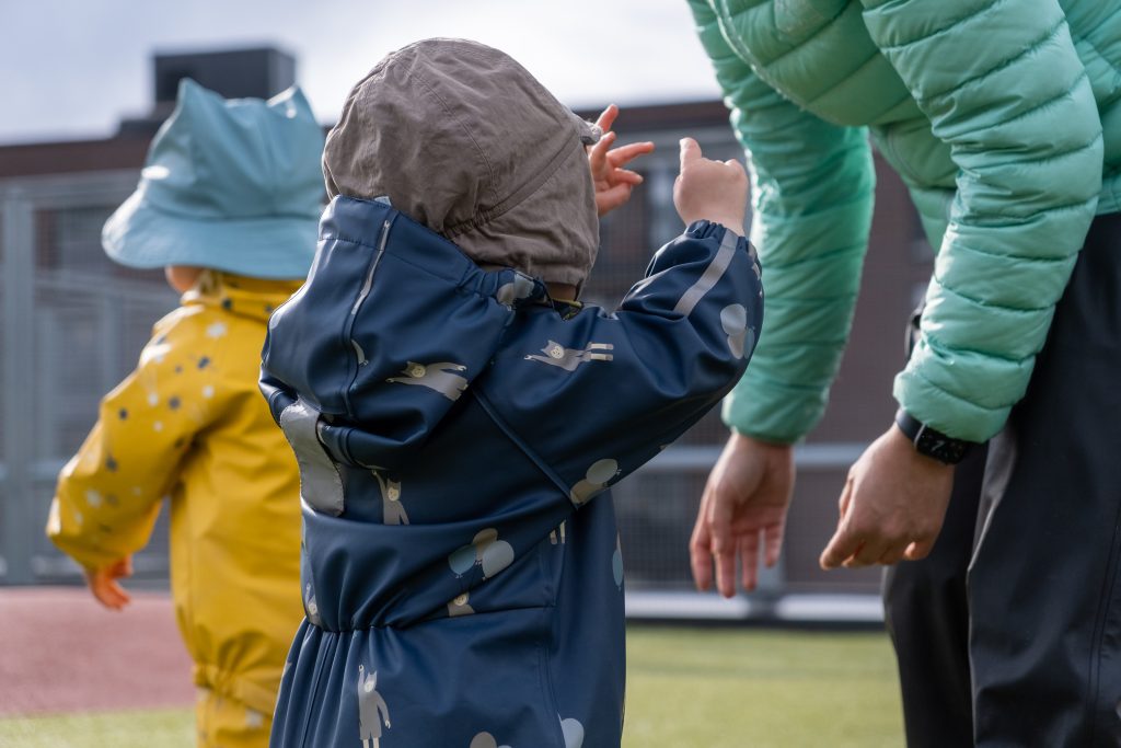 Bilde fra en banehage hvor et barn strekker seg mot en voksen. Man se også et annet barn i bakgrunnen. 