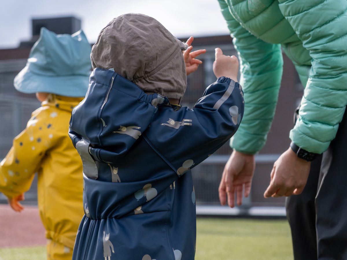Bilde fra en banehage hvor et barn strekker seg mot en voksen. Man se også et annet barn i bakgrunnen.