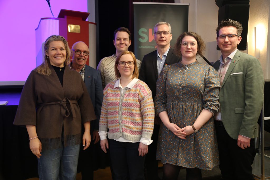 Gruppebilde av fylkesstyremedlemmene som var tilstede på årsmøtet. De står foran en SV-rollup med grønn bakgrunn og SVs logo. I venstre hjørne står det en talerstol. Bilde.