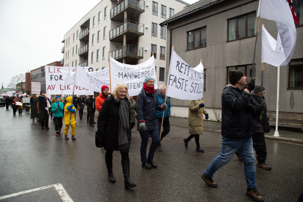Bildet viser et 1.maitog hvor de som går i toget holder paroler. I bakgrunnen ser man flere bygninger. Bilde