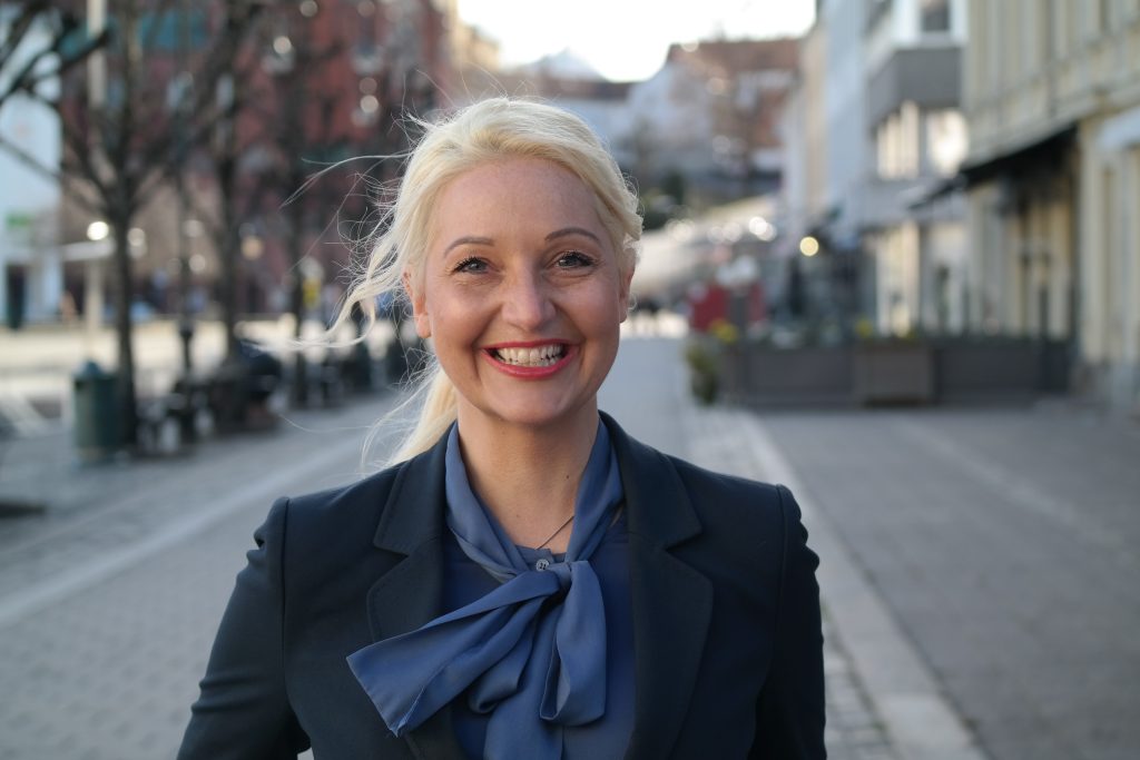 Portrettbilde av Stortingsrepresentant Mirell-Høyer-Berntsen som smiler. Hun står i en gate og i bakgrunnen kan man skimte bygninger og trær.